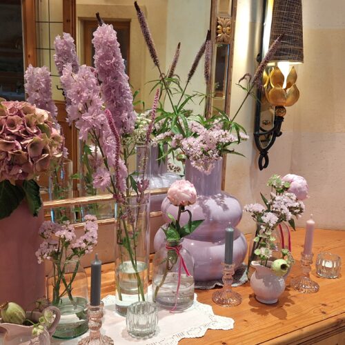 Verschiedene Gefäße mit rosa Deko und Blumen vor Spiegel auf Holzkommode