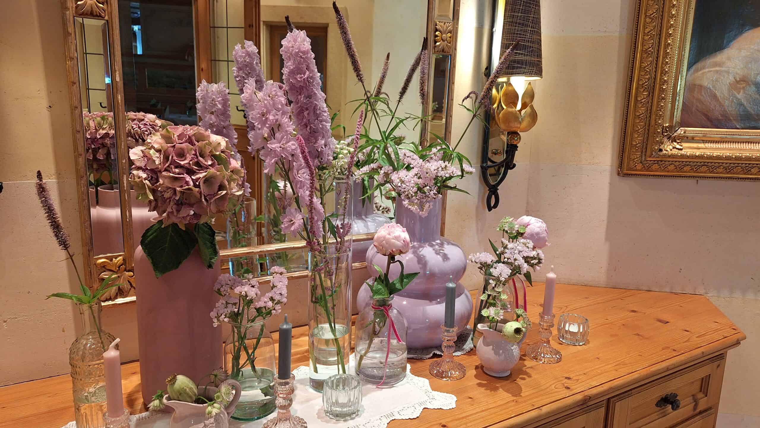 Verschiedene Gefäße mit rosa Deko und Blumen vor Spiegel auf Holzkommode